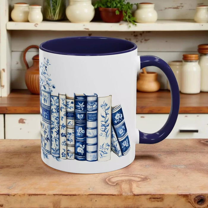 A white tote bag with a blue floral design and a stack of blue and white books, along with a matching blue and white mug, are displayed on a wooden surface.