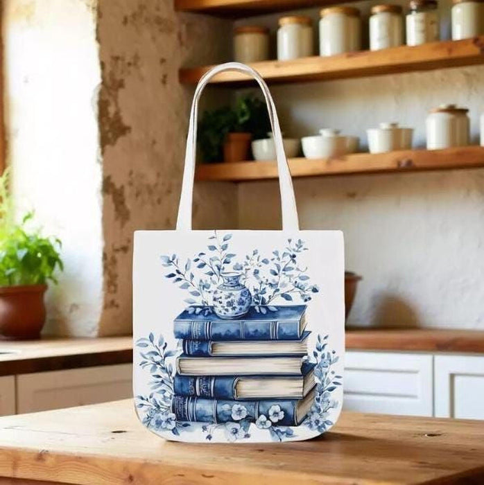 A white tote bag with a blue floral design and a stack of blue and white books, along with a matching blue and white mug, are displayed on a wooden surface.