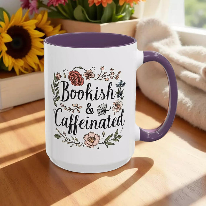 A white tote bag with a floral design and a purple handle, along with a white mug with a floral design and a purple handle, both sitting on a beige surface.