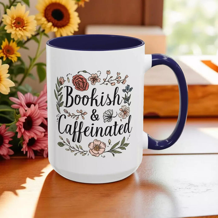 A white tote bag with a floral design and a purple handle, along with a white mug with a floral design and a purple handle, both sitting on a beige surface.